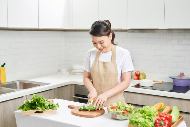 Jonge glimlachende huisvrouw bereidt gezonde maaltijd met groenten in lichte moderne keuken - Foto, afbeelding