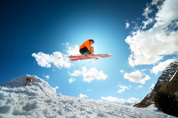 A young stylish man in sunglasses and a cap performs a trick in jumping with a kicker of snow against the blue sky and the sun on a sunny day. The concept of park skiing in winter or spring - Foto, afbeelding