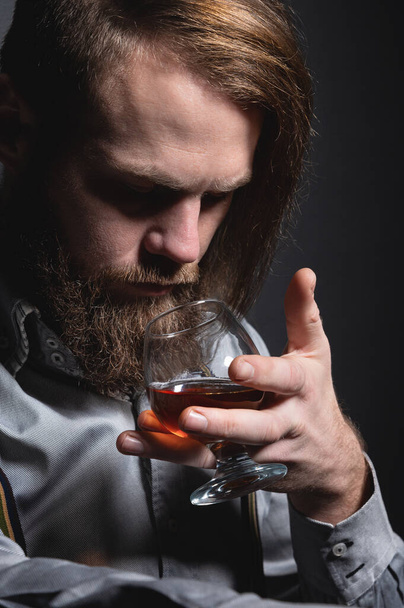 An attractive man with a long bang beard and a mustache sitting against the wall sniffs the scent of an alcoholic beverage in a glass that holds his hand. - Foto, afbeelding