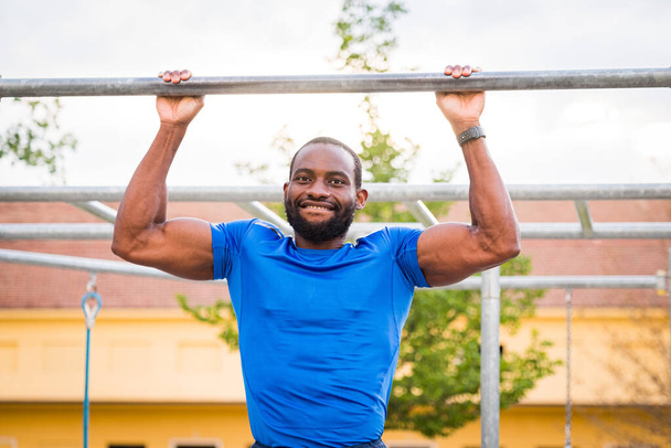 Αθλητής κάνει pull-up σε οριζόντια μπαρ - Fitness υπαίθρια στο πάρκο - Afro ή Αφρικής Αμερικανός άνθρωπος εξωτερική στην πόλη. Κάνε αθλητικές ασκήσεις. γυμναστήριο, υγεία, έννοια του τρόπου ζωής - Φωτογραφία, εικόνα