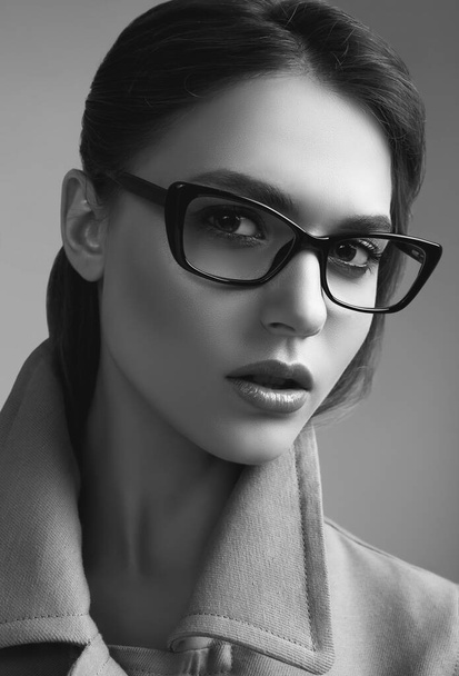 Портрет молодой красивой хипстерши в модном пальто и очках, позирующей на темно-сером фоне в студии
 - Фото, изображение