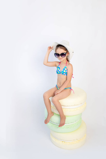 a little girl of 5-6 years old is sitting in a swimsuit with glasses in a hat on a white isolated background, a place for text - Foto, Imagen