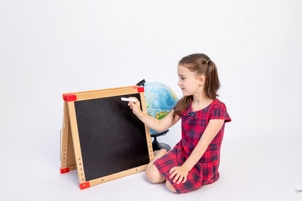 a little school girl of 7 years old sits in a red dress with a blackboard and writes with chalk on a white isolated background, place for text, September 1, knowledge Day - Photo, Image