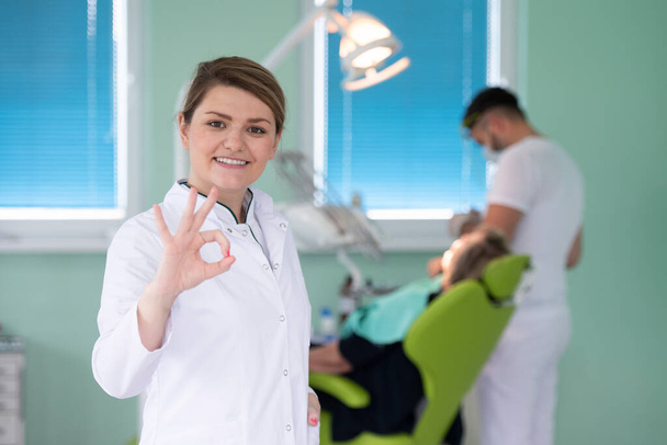 šťastný mladý ženský zubař ukazující ok znamení přes lékařskou kancelář zázemí, stomatologie a zdravotní koncepce - Fotografie, Obrázek
