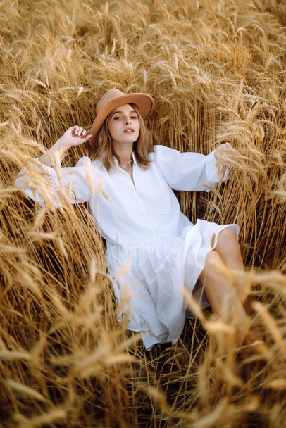 Стильна молода дівчина в літній білій сукні і капелюх позує на полі золотої пшениці. Мода, гламур, концепція способу життя
. - Фото, зображення