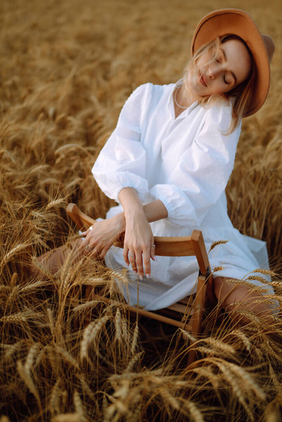 Stylish young girl in a summer white dress and hat posing in a golden wheat field. Fashion, glamour, lifestyle concept. - Foto, Imagen