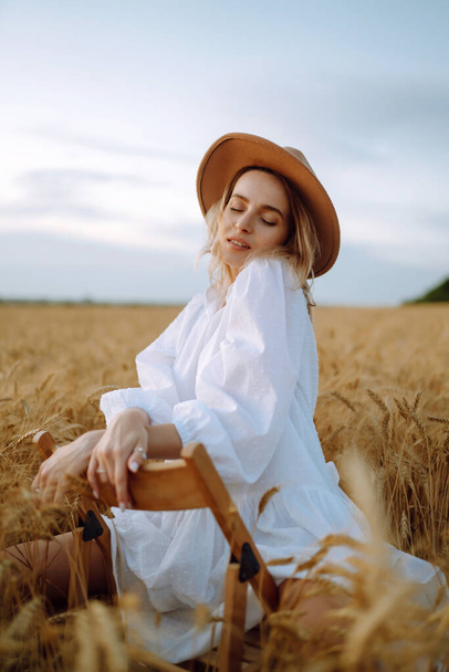 Stylish young girl in a summer white dress and hat posing in a golden wheat field. Fashion, glamour, lifestyle concept. - Foto, afbeelding