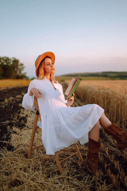 Stylish girl in white dress and hat with book in wheat field. Happy young woman on the sunset or sunrise in summer nature. Fashion and lifestyle concept. - Foto, afbeelding