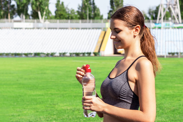 Beautiful woman is drinking water and listening to the music on headphones at the stadium. Girl is having a break after training. - Foto, Imagem
