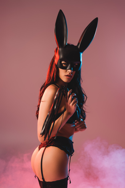 seductive girl in sexy underwear and mask with bunny ears holding flogging whip on pink with smoke - Photo, image