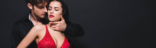 panoramatický záběr vášnivý muž dotýkat sexy dívka v červené podprsenka izolované na černé - Fotografie, Obrázek