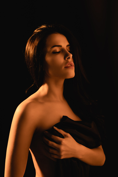 lighting on sensual woman with closed eyes wrapped in silk bed sheet isolated on black  - Photo, Image
