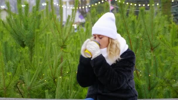 Mujer en sombrero blanco cálido, bufanda, manoplas se sienta a beber taza de papel de café
 - Imágenes, Vídeo