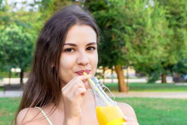 Attractive girl is drinking fresh juice through the straw outdoors. Pretty woman is holding a bottle of cold lemonade. - Photo, Image
