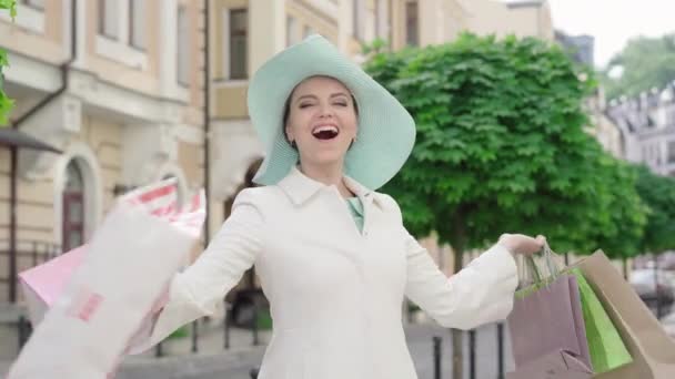 Middle shot of cheerful Caucasian stylish woman spinning with shopping bags and smiling at camera. Portrait of happy mid-adult lady in elegant hat and white coat rejoicing on sunny urban day. - Video