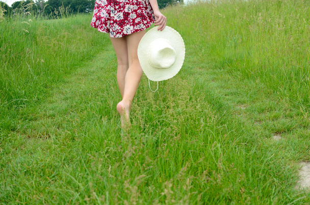 Barefoot girl walking on green grass. Female with colorful skirt, holding summer hat in hand. Summer in Poland, beautiful farmland in Lower Silesia. - Photo, Image