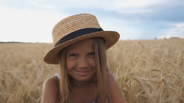 Gros plan de belle fille souriante en chapeau de paille regardant dans la caméra sur le fond de champ d'orge à la ferme biologique. Portrait d'heureux petit enfant aux cheveux blonds assis dans la prairie de blé - Séquence, vidéo