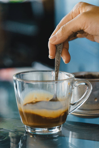 mani primo piano di una donna che mescola il caffè, caffè al latte caldo gocciolante in stile Vietnam. Concentrazione selettiva. Bere e concetto di viaggio - Foto, immagini