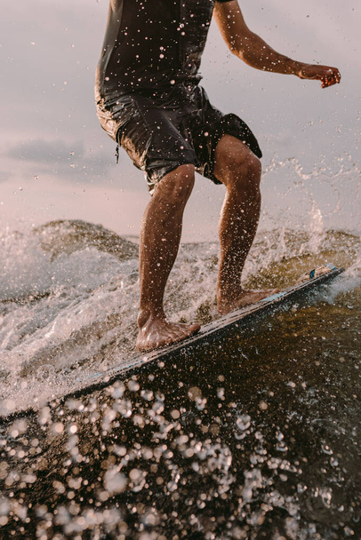 A sportive man is surfing on surfboard trails behind a boat. Wakesurfing on the river summertime leisure. Close-up of feet on the surfboard - Фото, зображення