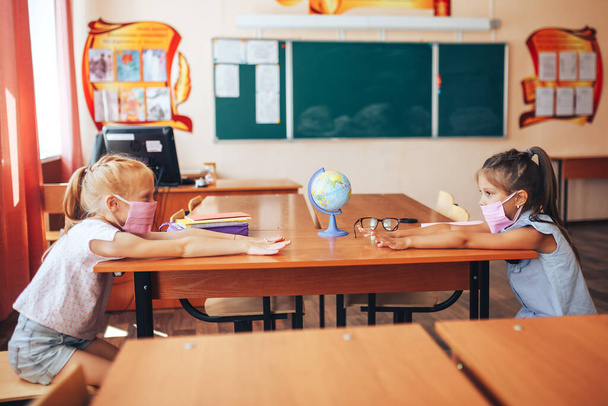 Két iskolás lány orvosi maszkban ül egy iskolaasztalnál, egymással szemben, csoportos foglalkozás, vissza az iskolába, gyerekeket tanítanak, társadalmi távolságot a járvány idején.. - Fotó, kép