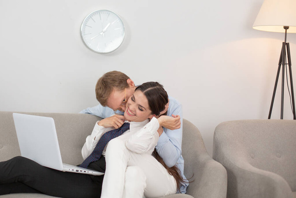 Couple resting on couch using laptop. Married couple having some relaxed time on sofa at home. Man kissing his girlfriend on neck while they sit on couch in living room with laptop - Photo, Image