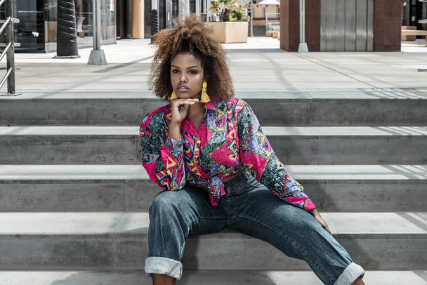 Afro saçlı, şık küpeli, renkli gömlekli ve kot pantolonlu, kentin caddesinde oturan ve kameraya bakan kendinden emin genç Afrikalı Amerikalı kadın. - Fotoğraf, Görsel