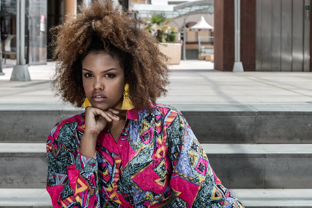 Σύγχρονη νεαρή Αφροαμερικανή γυναίκα με σγουρό χτένισμα φορώντας μοντέρνα μπλούζα με πολύχρωμο σχέδιο και κίτρινη φούντα σκουλαρίκια κοιτάζοντας την κάμερα, ενώ κάθεται στα σκαλιά κοντά στο κτίριο της πόλης - Φωτογραφία, εικόνα