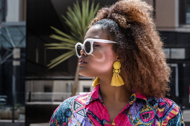 流行のサングラスでスタイリッシュな若いアフリカ系アメリカ人女性と街の通りに立ってカラフルなブラウスを身に着けている明るい黄色のタッセルイヤリングを自信を持って離れて見て - 写真・画像