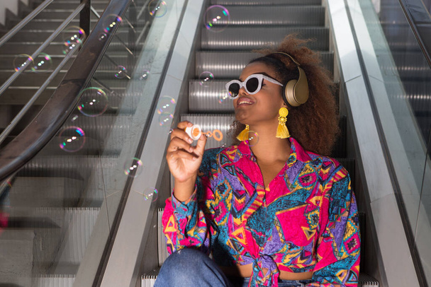 Angle bas de femme gaie dans une usure lumineuse et bijouterie soufflant des bulles fragiles et transparentes tout en étant assis sur des escaliers mobiles dans un casque sans fil - Photo, image