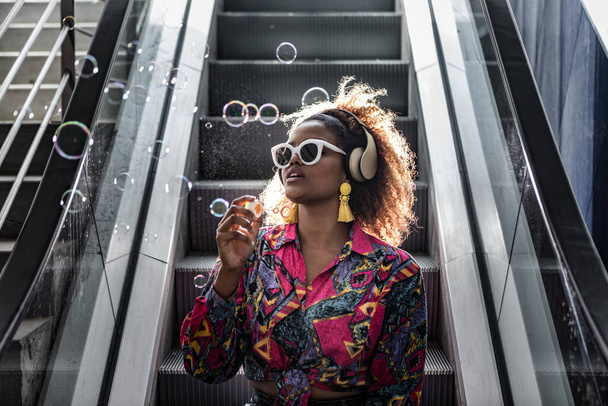 Mujer afroamericana en gafas de sol y blusa colorida sentada en una escalera móvil con la boca abierta mientras sopla burbujas en la espalda iluminada
 - Foto, Imagen