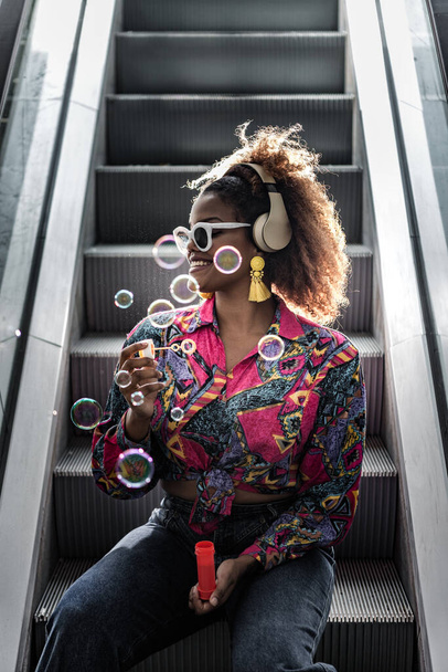 Smiling African American female in wireless headset and yellow earrings blowing bubbles while sitting on moving staircase step in back lit and looking away - Photo, Image