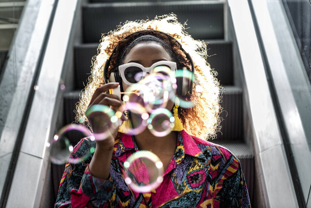 Anonymous African American female in headphones and colorful blouse blowing transparent bubbles while sitting on moving staircase in back lit - Photo, Image