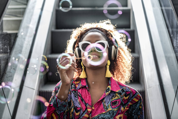 Content African American female in yellow earrings and modern headset blowing fragile bubbles while sitting on moving staircase in back lit - Photo, Image