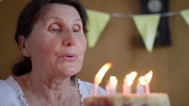happy birthday, elderly woman enjoys holiday and blows out burning candles on sweet birthday cake, grandmother celebrates anniversary and holds birthday cake in her hands, close-up - 映像、動画