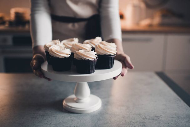Frau backte leckere Schoko-Cupcakes mit geschlagenem Frischkäse, die in Kithen-Großaufnahme auf dem Tisch standen. Arbeiten zu Hause.  - Foto, Bild