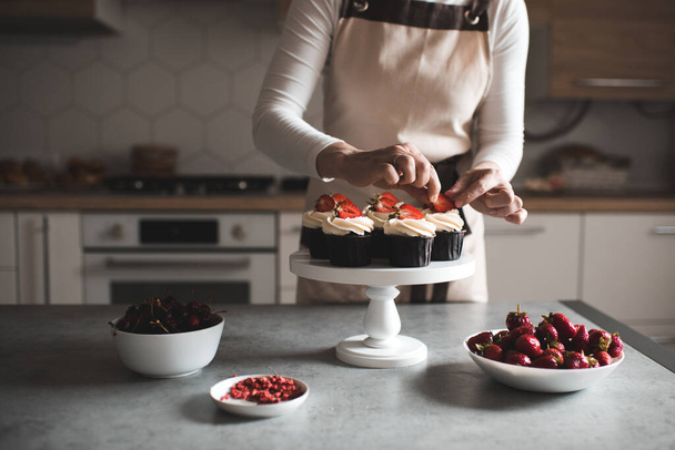 Молодая женщина украшает кексы фруктами и сливочным сыром крупным планом на кухне. Работаю дома. Селективный фокус.  - Фото, изображение