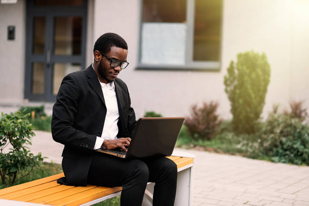 Przystojny młody, afrykański Amerykanin pracujący na laptopie, siedzący na zewnątrz na ławce. - Zdjęcie, obraz