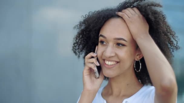 若い陽気なアフリカ系アメリカ人の女性の近くに歩くと、華やかに前のビッグガラスビル街の通りで電話で話している。モバイルネットワーク上の友達とコミュニケーションをとり - 映像、動画