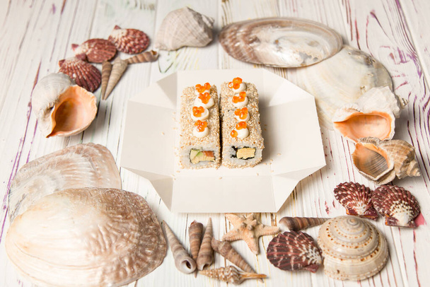 Πλάγια άποψη των ασιατικών σούσι στο πακέτο παράδοσης κουτί σε ξύλινο φόντο με θαλασσινά όστρακα και αστέρια. Roll με σολομό, αβοκάντο, σουσάμι, ιαπωνική μαγιονέζα και κόκκινο χαβιάρι στην κορυφή. - Φωτογραφία, εικόνα