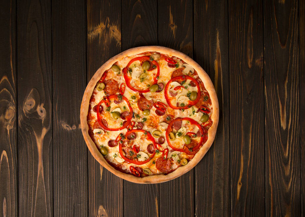 Vista superior de pizza con pimiento, aceitunas verdes, salchichas ahumadas, queso mozzarella, pepperoni, salami, chile, champiñones sobre fondo de madera. Sabrosa pizza sin cortar
 - Foto, Imagen