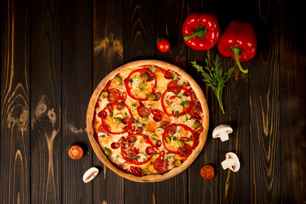 Vue de dessus de pizza au poivron, olives vertes, saucisses fumées, fromage mozzarella, pepperoni, salami, piment, champignons sur fond bois. Savoureuse pizza non coupée avec des ingrédients et des herbes - Photo, image
