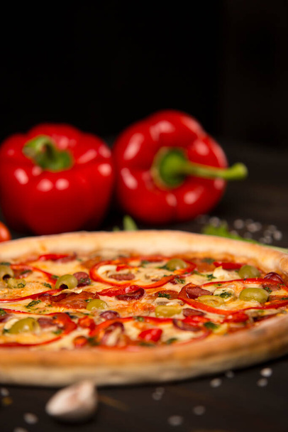 Side view of pizza with bell pepper, green olives, smoked sausages, mozzarella cheese, pepperoni, salami, chili pepper, champignons on wooden board with two bell peppers on background. uncut pizza - Foto, afbeelding