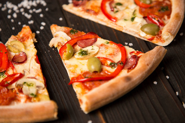 Slice of delicious pizza. Side view of pizza with bell pepper, olives, sausages, mozzarella cheese, pepperoni, salami, chili pepper, champignons on wooden background with sea salt. Close up of piece - Фото, изображение