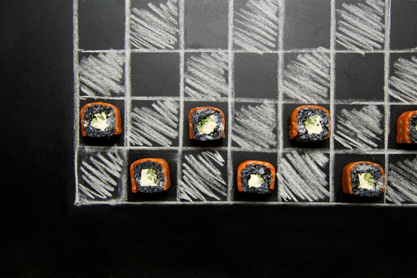 Philadelphia Sushi-Rollenstücke mit schwarzem Reis auf dunklem Kreidegrund mit Kreidezeichnung. Japanisches Sushi mit Lachs, Gurken und Frischkäse auf handgezeichnetem Schachbrett. - Foto, Bild