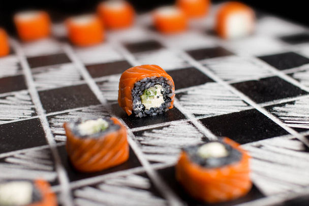 黒板に黒と白のフィラデルフィアの寿司ロールピースでチェスをする。手描きのチェスボードにサーモン、キュウリ、クリームチーズの和え寿司。黒米寿司 - 写真・画像