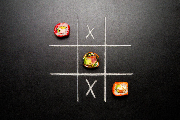 Ręcznie rysowane Tic Tac Toe gra z japońskim sushi roll sztuk zamiast zera na ciemnym tle tablicy. Azjatyckie bułki z łososia, kraba śnieżnego, awokado i zdrowej bułki z ogórkiem zamiast ryżu. - Zdjęcie, obraz