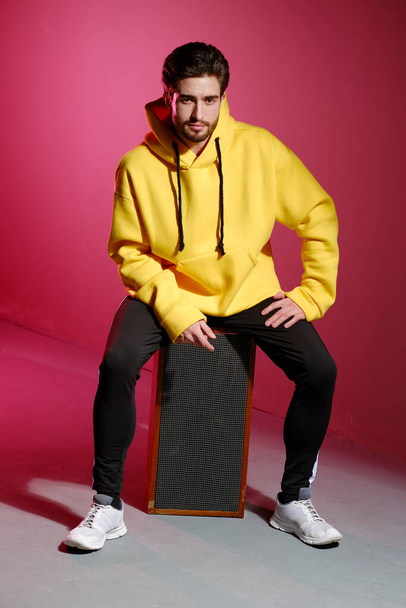 A young man of 25-30 years old in a yellow sweatshirt sitting on an old music column on pink wall background and showing different emotion. - Fotografie, Obrázek