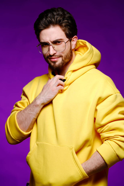 Młody mężczyzna od 25-30 lat w okularach i żółta bluza emocjonalnie pozuje na fioletowym tle..  - Zdjęcie, obraz