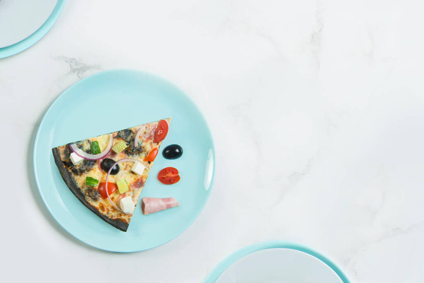 Slice of meat pizza with vegetables. Dish with cherry tomatoes, cucumber, Feta cheese, ham, black olives and Spanish onion on black dough. ingredients on pastel serving plate, marble background. - Foto, Bild