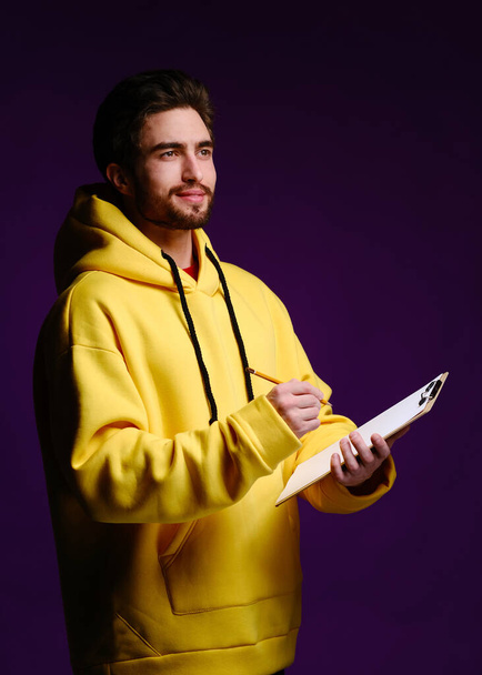 A young man of 25-30 years in glasses and a yellow sweatshirt holding a clipboard with clip in one hand and a pencil in the other - Photo, Image